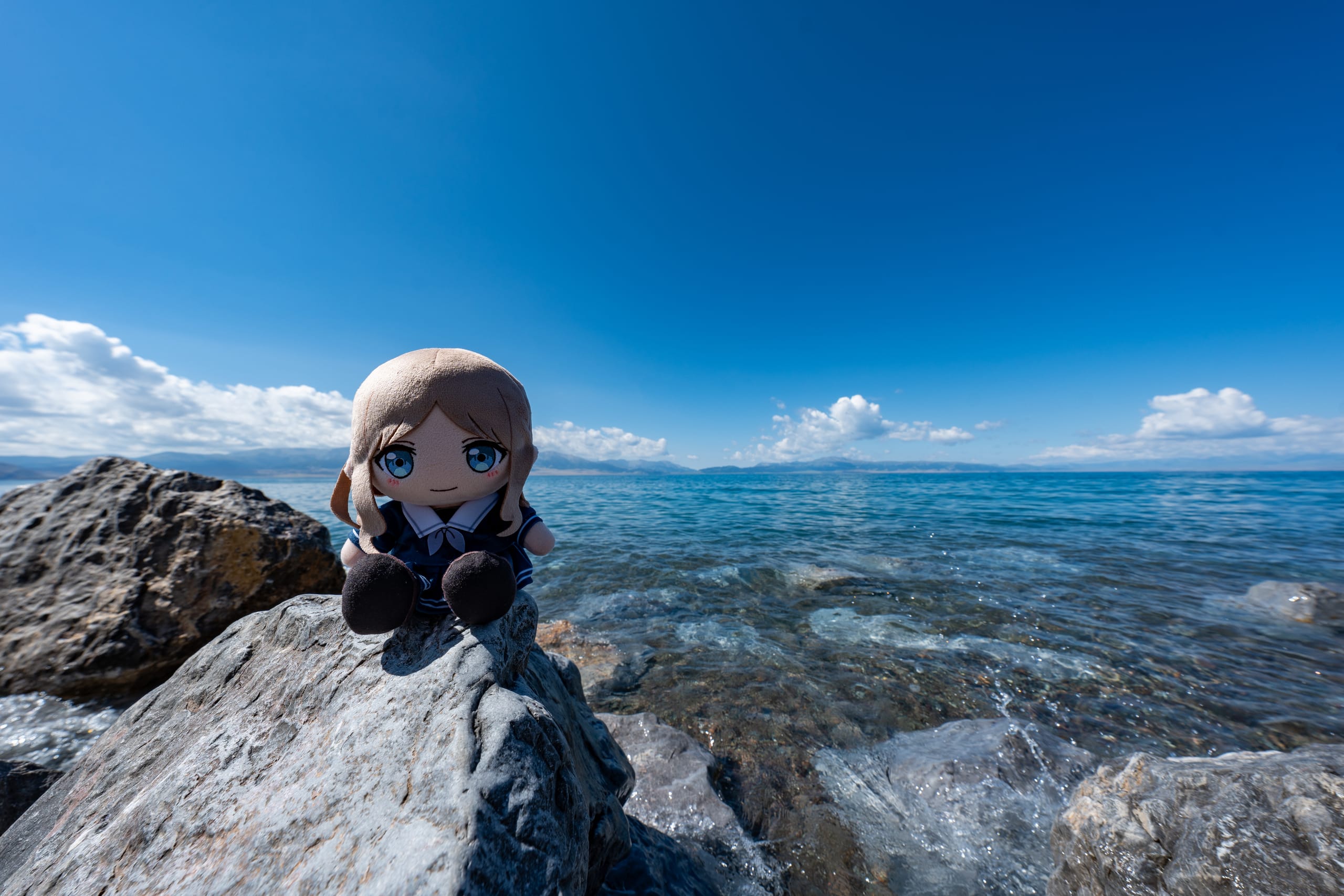 soyorin玩偶坐在赛里木湖岸的岩石上，背后是蓝色湖水与远山。