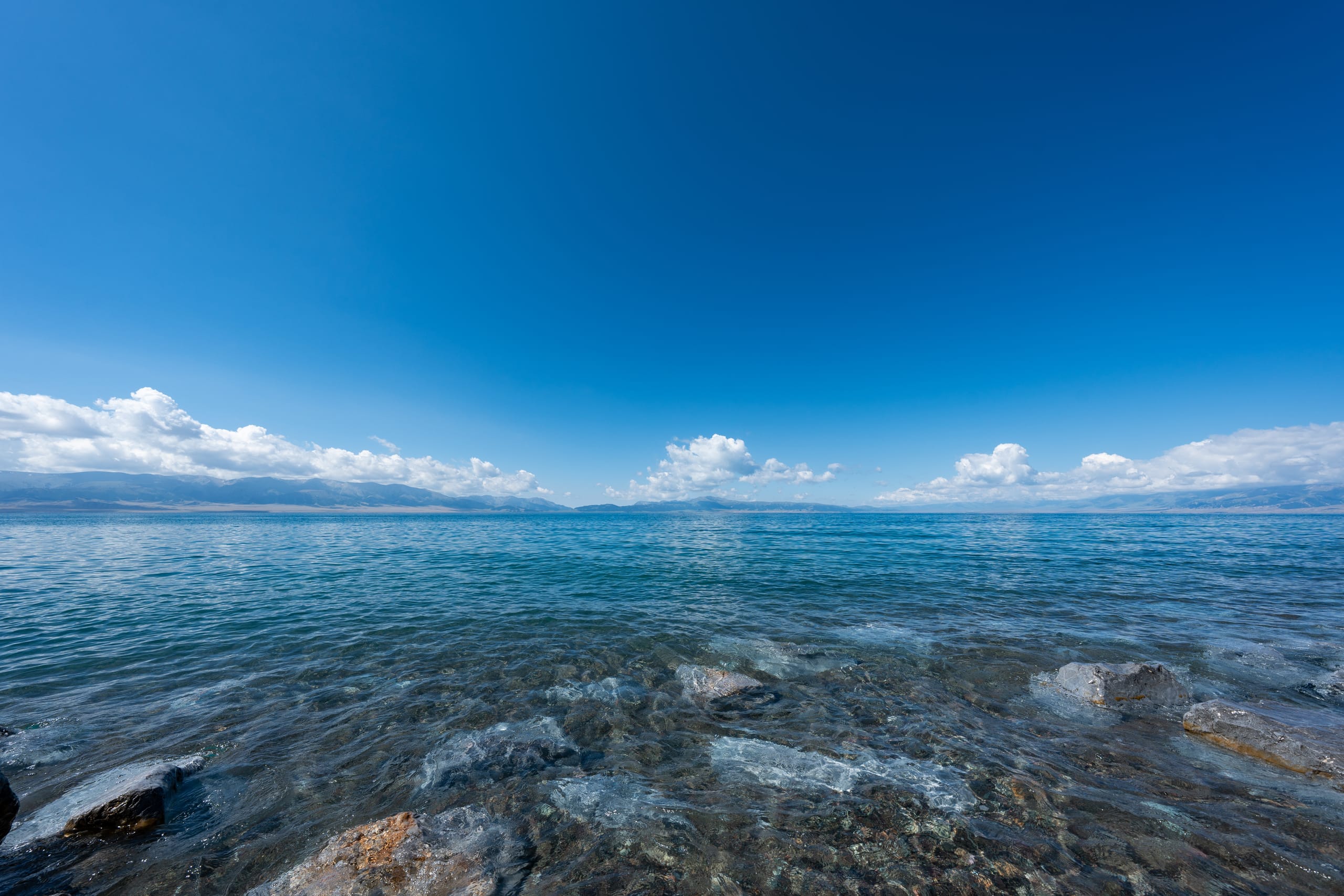 赛里木湖清澈水浪涌向岩石岸边，远处雪山位于云层下。