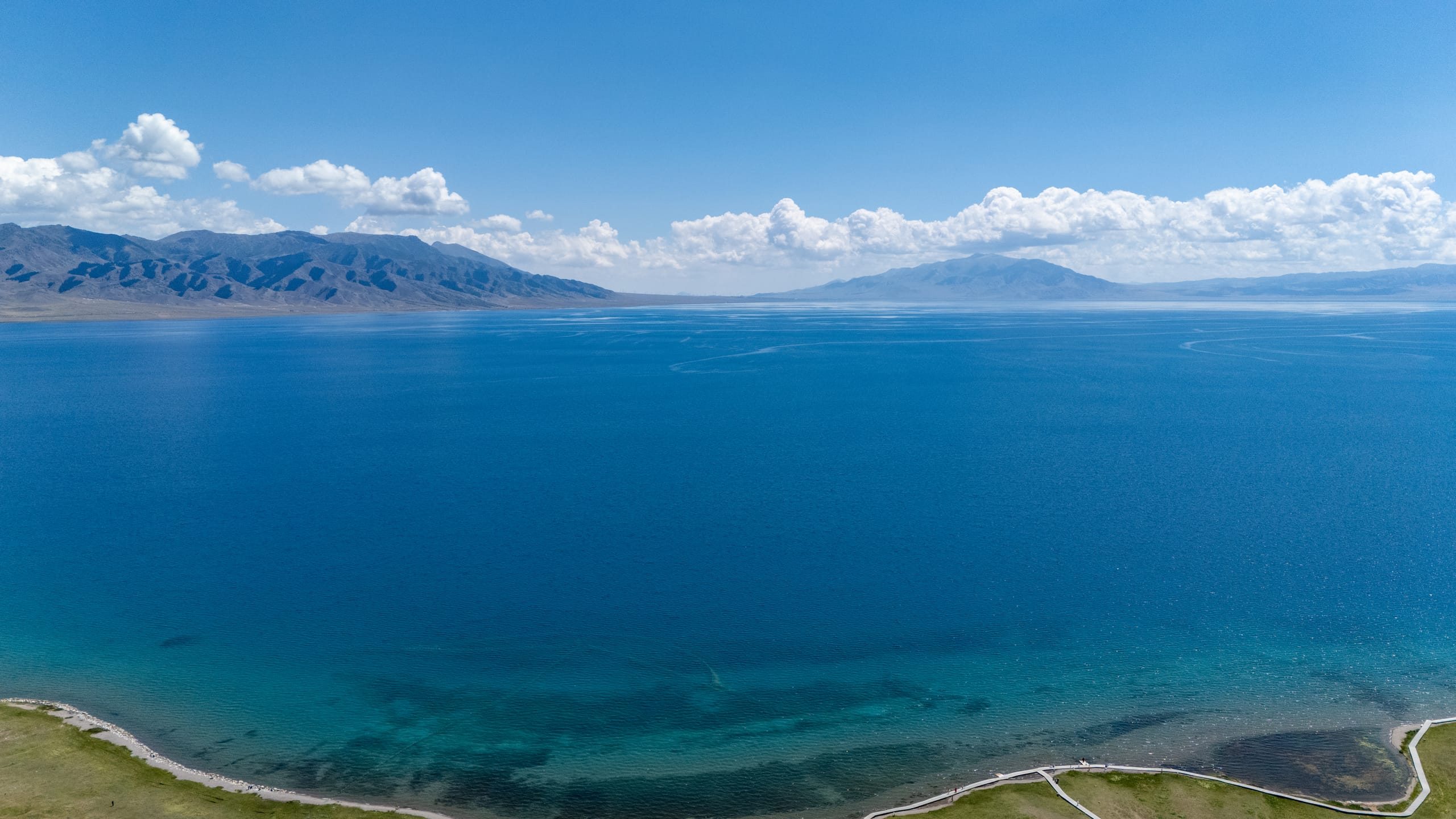 航拍赛里木湖近岸，透明浅水、弧形道路与深蓝湖面向远山延伸。