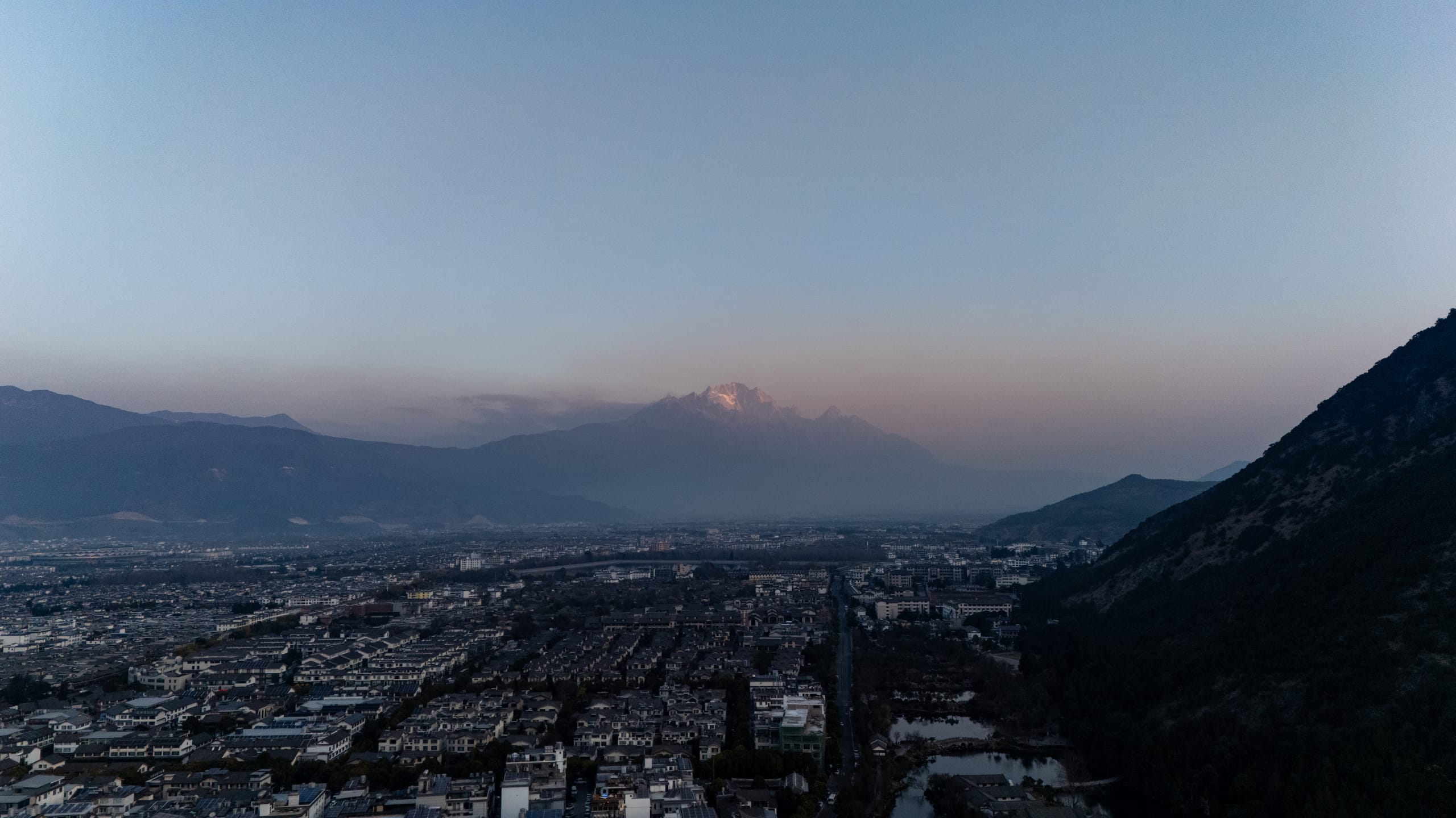 清晨航拍丽江古城，城镇铺展在暗色山谷中，远处玉龙雪山隐在薄雾里。
