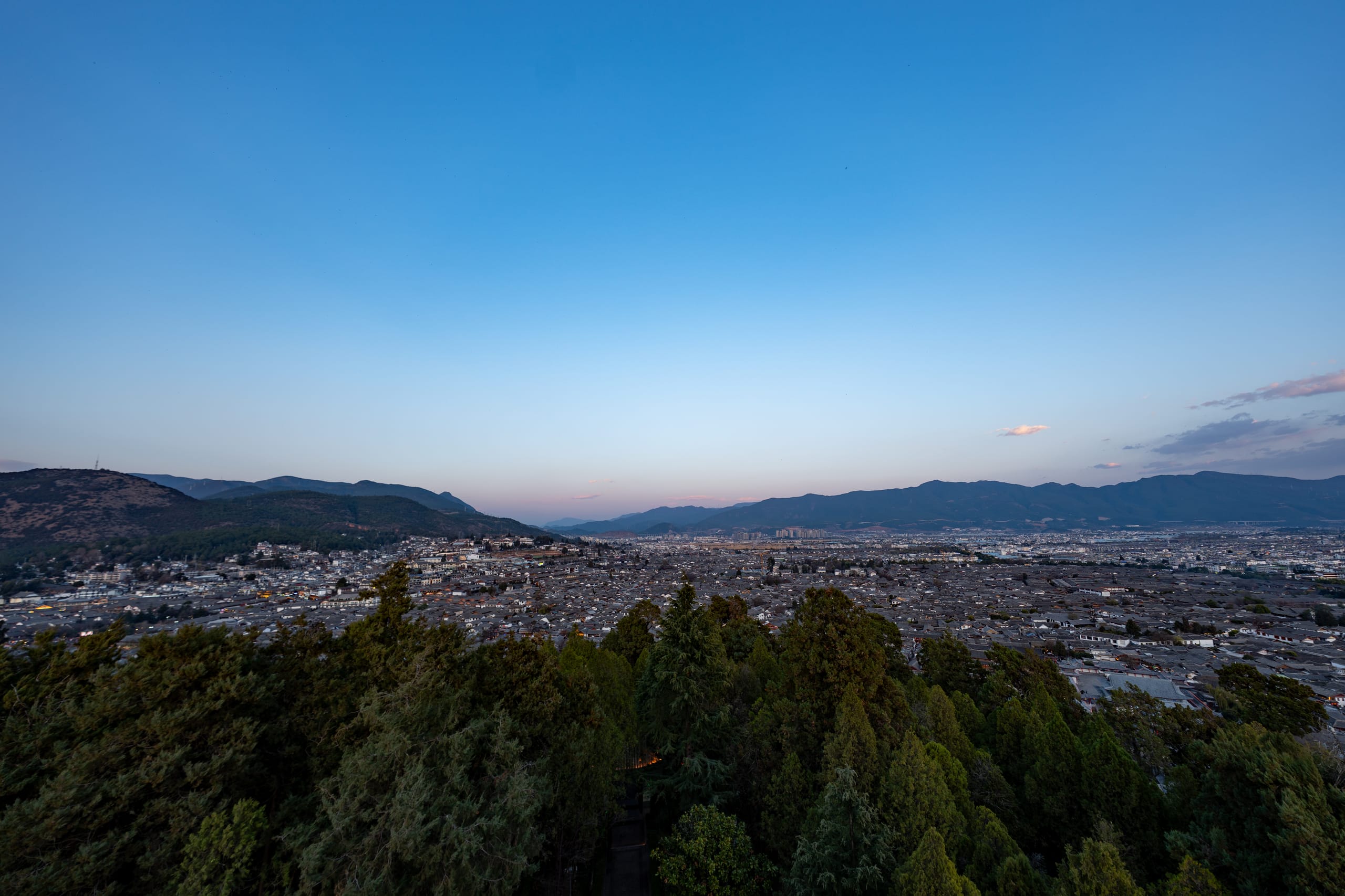丽江古城与周边城区铺展至远处群山，前景为层叠树木。