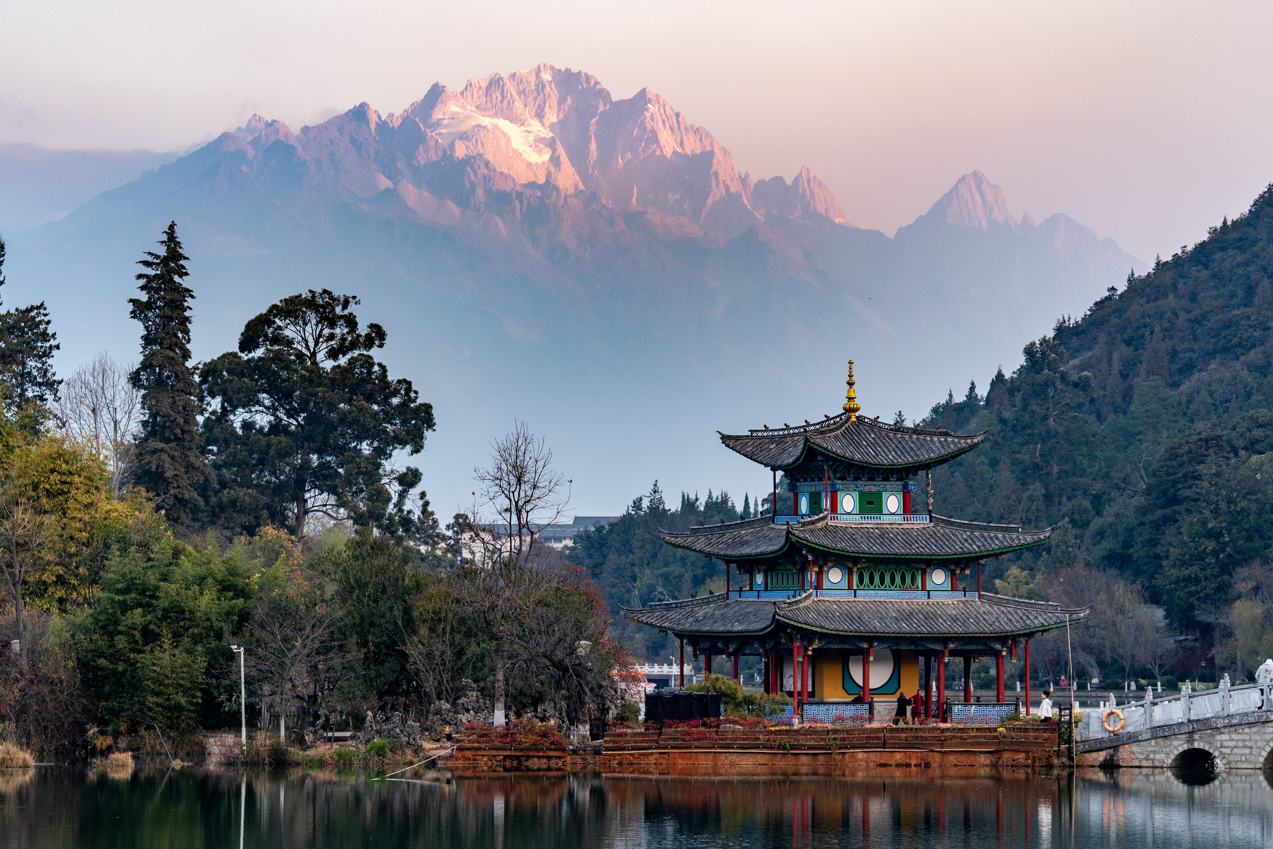 黑龙潭亭阁倒映在水面，晨光照亮其后的玉龙雪山。