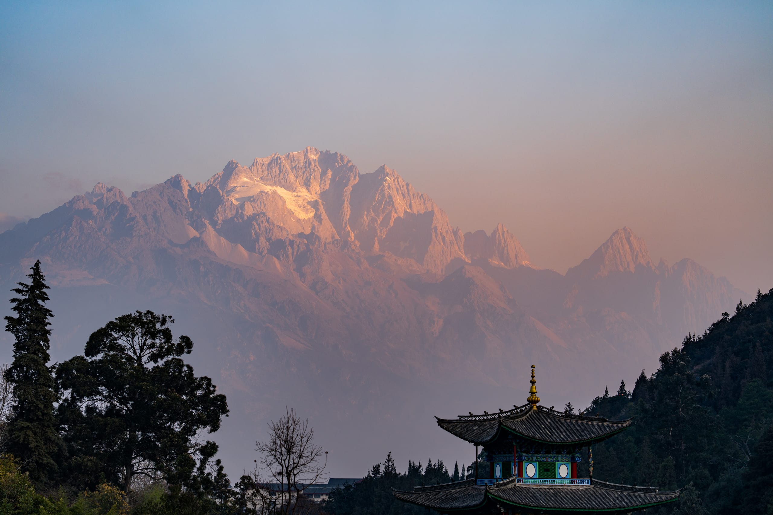 清晨的黑龙潭亭阁与玉龙雪山，淡金色阳光穿过薄雾落在山峰上。