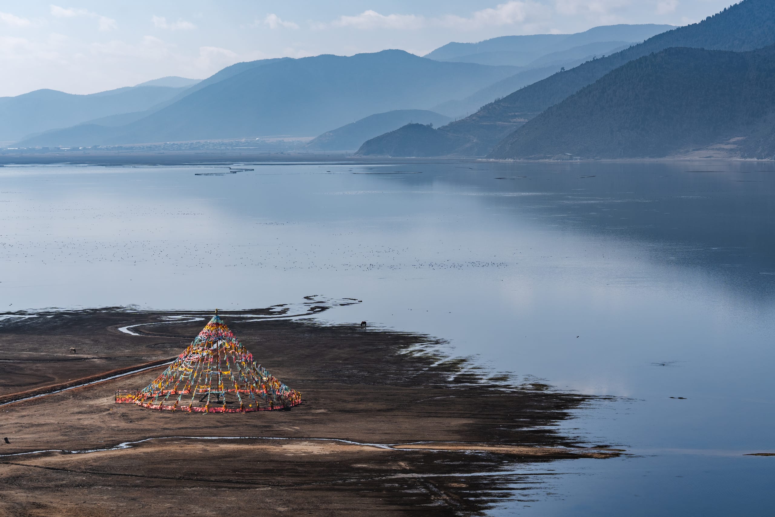 高处俯瞰纳帕海浅滩与湖水，彩色经幡架立在岸边，远山层叠。