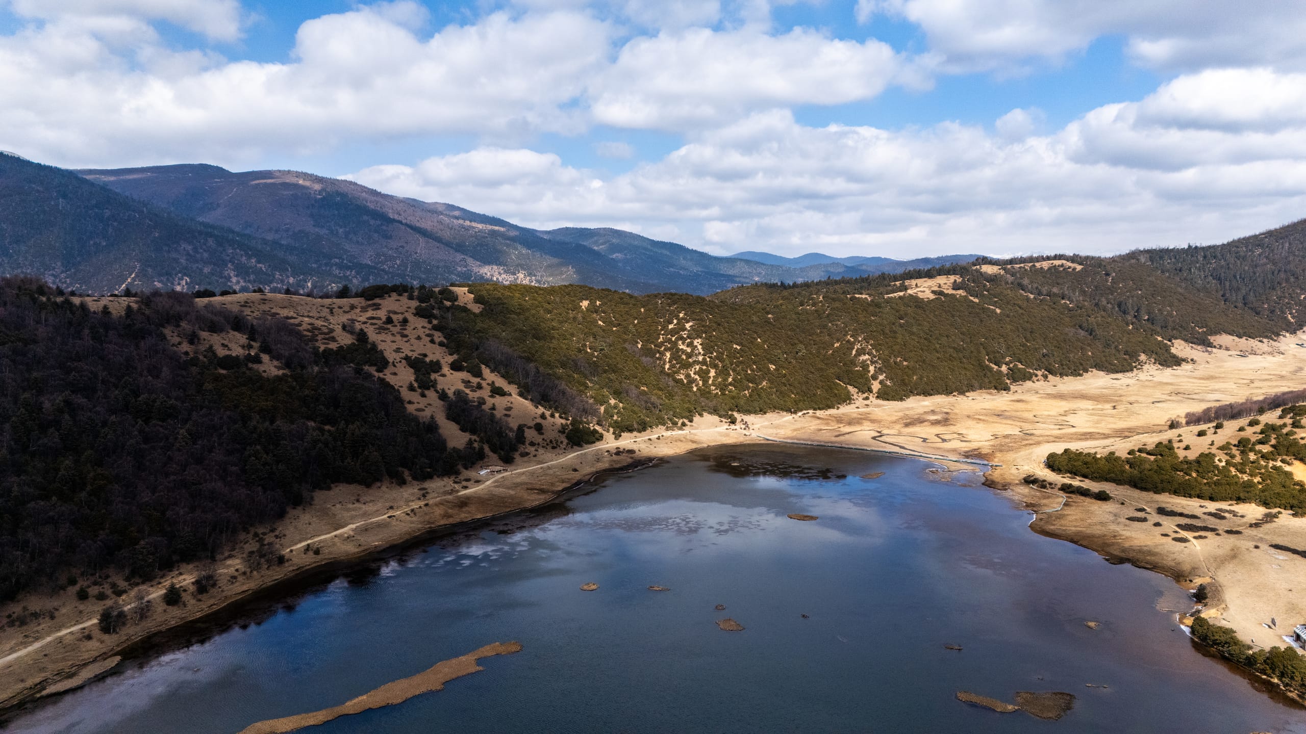 空中侧望属都湖弯曲湖岸，金色草地与深色山林环绕蓝色水面。