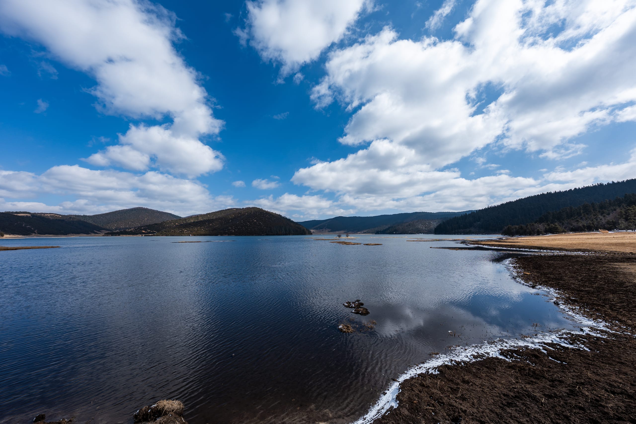 属都湖全景，弯曲湖岸、远山和蓝天白云倒映在深色水面。