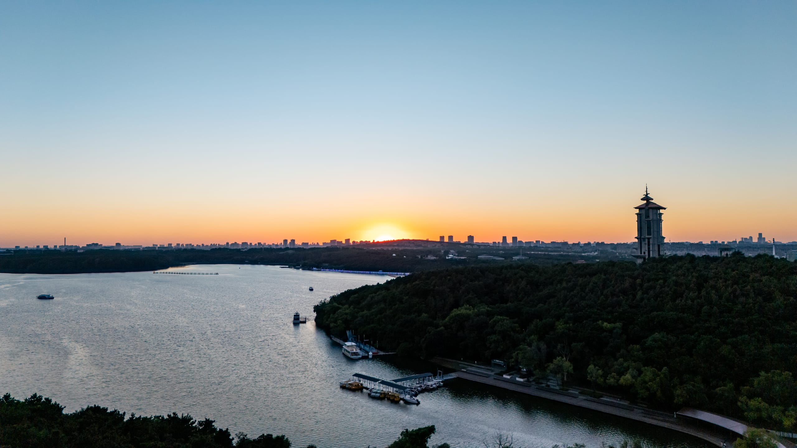 净月潭暮色全景，夕阳、岸边亭楼与城市天际线沿水面展开。