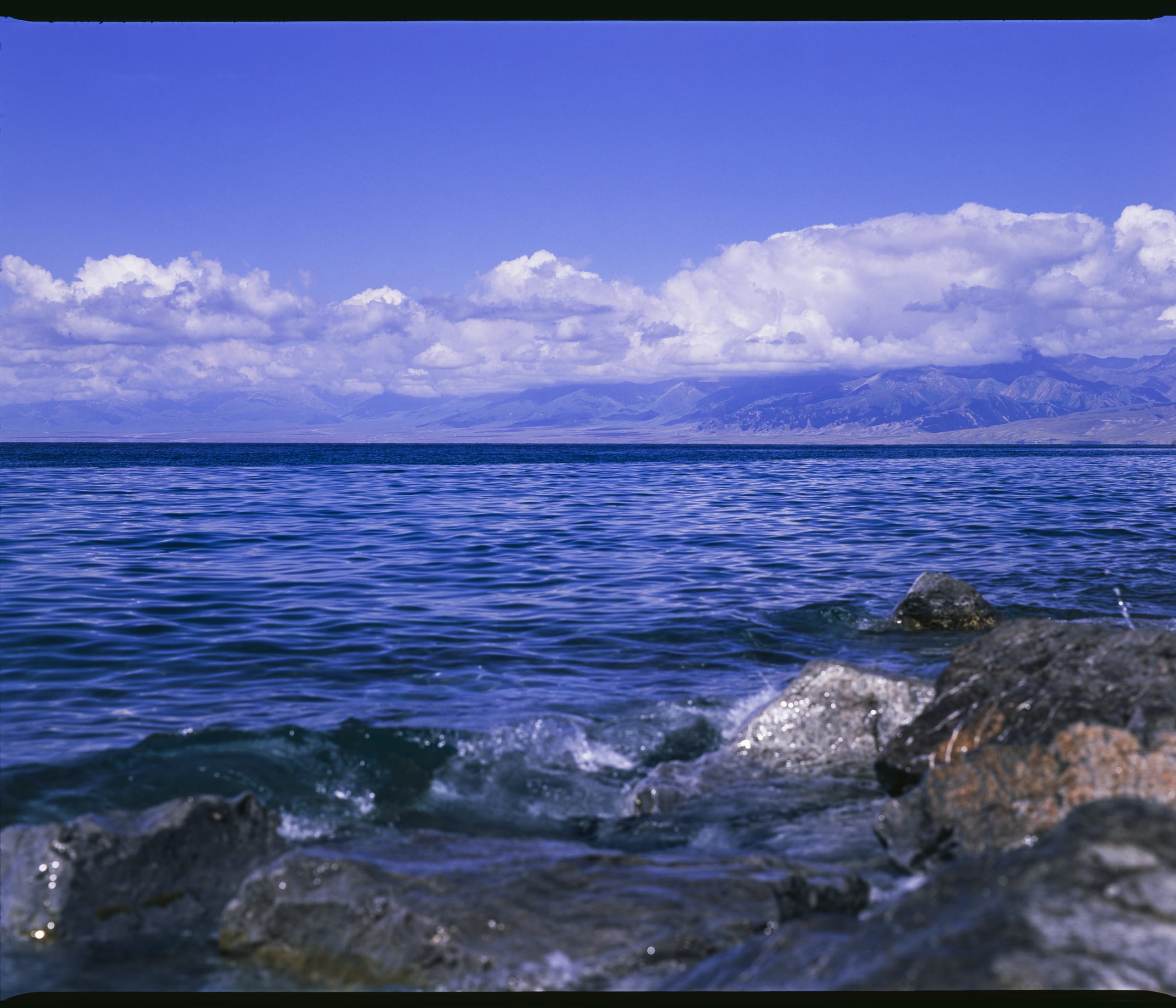 岩石岸边的湖水泛起波浪，远处山脉上方铺展着白云